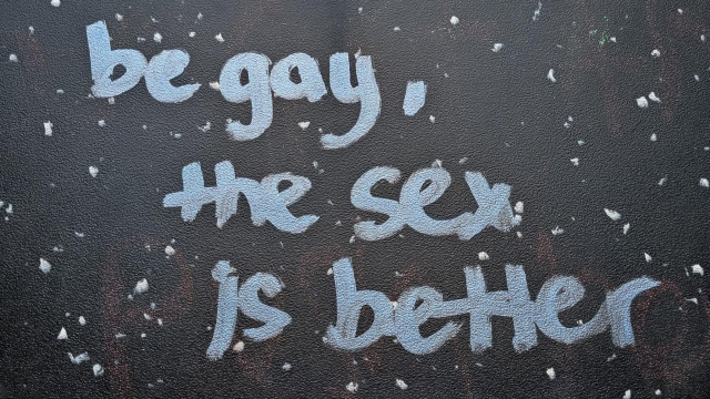 A speckled black restroom stall wall. Light blueish-white graffiti reads "be gay, the sex is better".