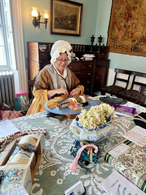 White woman in 18th century American costume (yellow and green striped bedgown, white linen ruffled hat stuffed with cotton to look poofy). I have a black gauze shawl with black lace trim on the bottom over top of my outfit. I made the lace but not the gauze part. I am seated at a dining table.

In front of me is my lace pillow with dozens of bobbins making more black silk lace. My colleague's pillow is off to the left a smidge. We are at a dining room table in an 18th century house with period furniture and tapestry and print stuff on the walls. 

The dining room table has a filet lace large table cloth on it. It has a tureen filled with flowers in the center.