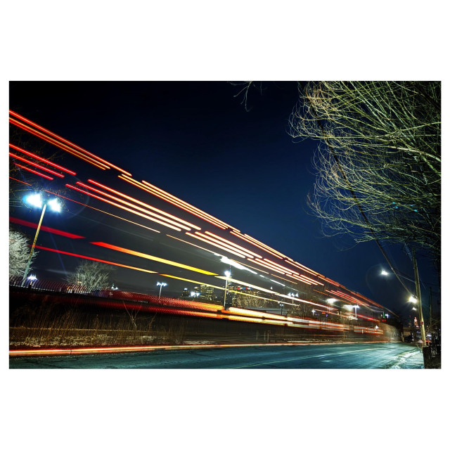 Nighttime long-exposure photo of red and yellow light streaks from the taillights of a passing truck, on a road by the Brooklyn waterfront. Through the streaks we can see glowing streetlights and a glimpse of the lit-up windows of the lower Manhattan skyline. At right are the leafless branches of a tree and above is a clear sky, deep blue in the early evening.