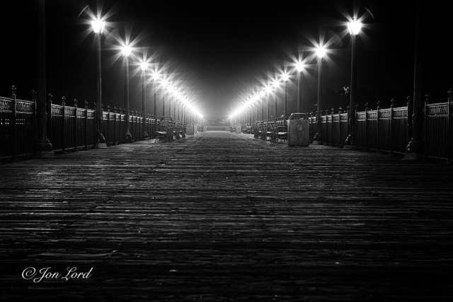 This is a night time, grungy, black and white photo in landscape format looking along a wooden pier lined with street lights that converge into the distance. The Embarcadero, San Francisco (2012).

The camera is about half a metre above the weathered and rough wooden planks that lay at right angles to the lens. The lighting above exaggerates the knurled and uneven texture of the well worn wooden surface. At the edges of the pier are two parallel rows of ornate, black metal railings. Spaced every three or four metres are dark metal street lights that are about three or so metres tall. Each street light is topped with a lantern that radiates outwards eight petal-like rays. Arranged along the edges are numerous park benches plus the odd large litter bin. Both the railings and street lights while in reality are parallel: in the image converge together with increasing distance. Above and to the sides of the scene is a pitch black and featureless sky. 

