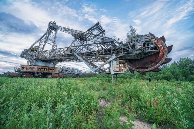 A massive, rusty bucket-wheel excavator, an industrial relic, stands abandoned in a vibrant green field under a cloudy sky. Its sheer scale is emphasized by a distant person.