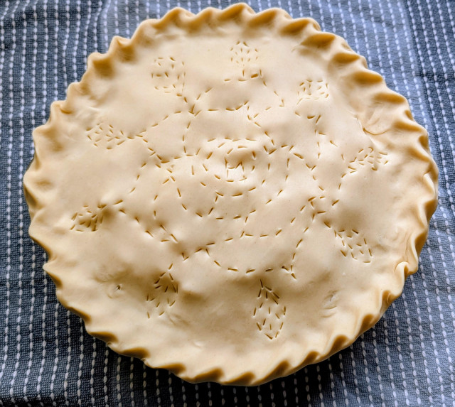 An unbaked apple pie with a vent design in the shape of a rose with leaves around the edges. 