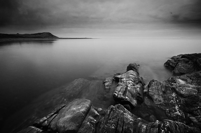 Atmospheric black and white photo of rocks in a calm sea, with a headland in the middle distance and dark clouds above.