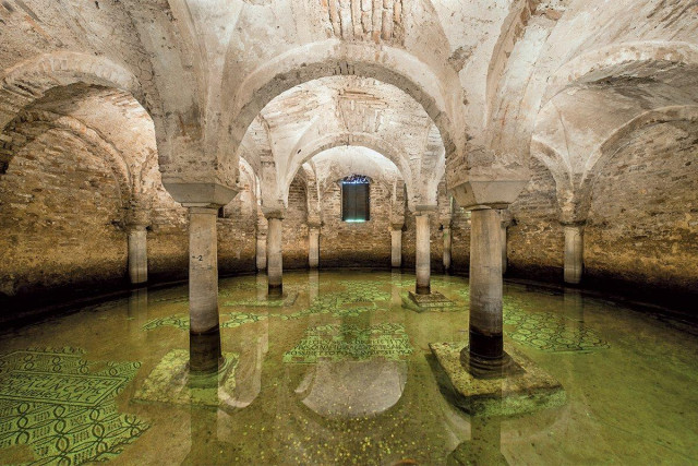 An interior view of the ancient, flooded crypt of the Basilica di San Francesco in Ravenna. Shallow, clear green water covers the floor, where intricate, mosaic patterns are visible beneath the surface. Stone columns with vaulted arches stand in the water, their bases partially submerged.
