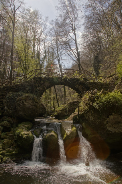The image depicts a serene natural scene featuring a stone arch bridge with a wooden railing, spanning a small waterfall. The bridge is covered in moss and surrounded by lush greenery, including trees with sparse leaves, indicating early spring. The waterfall cascades over moss-covered rocks into a pool below, with sunlight filtering through the trees, creating a lens flare effect. The scene is tranquil, with the water flowing gently over the rocks, and the overall atmosphere is one of peacefulness and natural beauty.