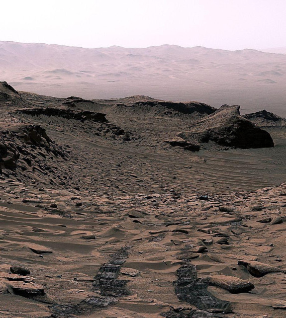 Photo of the Martian landscape, looking back downhill at the tracks of the Curiosity rover. Sands cover the area, with several rough outcrops lifting up to either side of a gully. The distant hills are hazy.