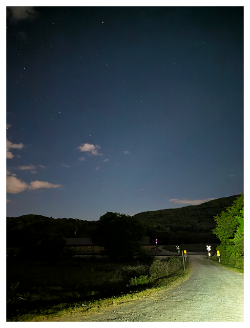 nighttime rural scene shows a gravel road crossing railroad tracks, illuminated by headlights. rees and hills are visible under a dark, starry sky with a few clouds.