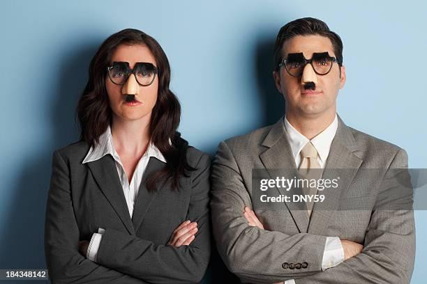 Color photograph.
A woman and a man standing next to one another dressed in business attire with their arms crossed in front of them.
Both people are wearing Groucho glasses: Dark eyeglass frames with heavy black eyebrows, a large nose, and a black mustache.
Photo has a grey rectangle superimposed on it that reads, "getttyimages. Credit: DNY59."
