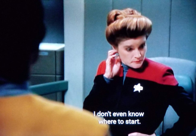 Star Trek Voyager scene. Captain Janeway, with her hair up and bouyant this season is standing in her ready room. Someone is facing her but we can only see the back of them half off-screen. Janeway has one hand on the hip and the other hand raised up to her ear and is looking down like she's irritated or thinking hard. Closed caption reads, "I don't even know where to start."