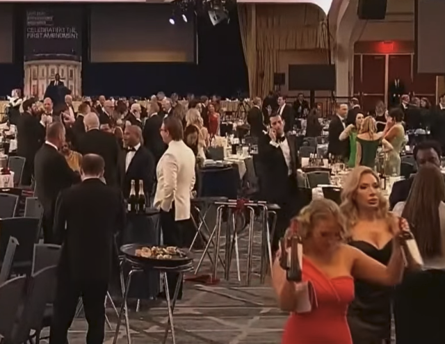 A woman in a red dress leaving the White House Correspondents' Dinner with her arms raised holding two bottles of wine.