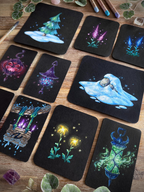 A photo of several small original paintings on black paper, placed on a wooden table and surrounded by a paint brush, coloured pencils, leaves, and crystals
 The paintings feature: a snowy tree, glowing pink flowers, glowing blue flowers, a demonic red lantern,  a witchy purple lantern, a snowy rock, a hanging sign with glowing mushrooms on it, glowing yellow flowers, and a spectral green lantern. 