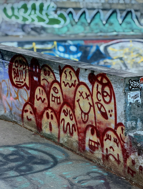 A color, wide-angle photograph in portrait orientation that captures a concrete ledge covered in vibrant street art at a skate park in the Bronx, New York. The primary focus is a cluster of approximately thirteen figures spray-painted in a deep rust-red and yellow hue onto the side of a weathered grey concrete wall. Each figure is a simple, rounded oblong shape, yet they are individually characterized by unique, expressive facial features. Some display wide-eyed shock, others look grumpy or sleepy, one has "X" marks for eyes with its tongue sticking out, and another features a single cyclopean eye. To the far left, a speech bubble contains the hand-lettered words "ODD BALL!" in the same red paint. The concrete itself shows signs of age with mottled textures and small cracks, adding a gritty, urban feel to the composition. The background and foreground are softly blurred, creating a shallow depth of field that emphasizes the central red figures. In the background, larger, more complex graffiti tags in shades of emerald green, bright blue, and white cover distant walls, though their details are out of focus. On the right edge of the main concrete block, a small black-and-white sticker is visible with the words "CLEARLY FADED". The lighting is bright and even on an overcast morning that allows the rich red tones of the figures to stand out against the cooler greys and blues of the park.