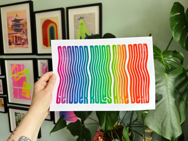 A hand holds a colorful print with the text "GENDER ROLES ARE DEAD" in a wavy design, featuring a gradient of vibrant rainbow gradient 