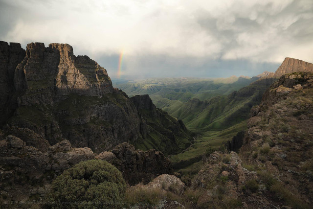 A view from high on the Drakensberg escarpment looking down through a narrow gorge into a broad, lush green valley far below, with a partial rainbow arcing above the sunlit cliff face on the left under a dramatic stormy sky. Dark basalt towers and rocky outcrops frame the foreground on both sides, while the verdant valley floor and rolling green foothills stretch into the distance beneath a break in the clouds. The contrast between the brooding storm overhead and the sunlit landscape below gives the scene a powerful, charged atmosphere.