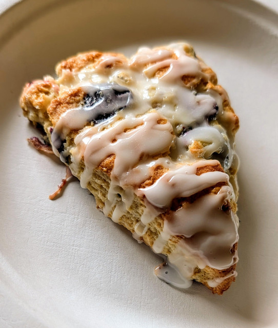 Lemon blueberry scone with drizzled lemon frosting on a white paper plate. 