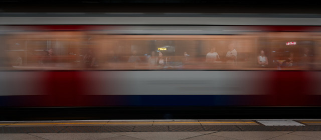 A subway train rushes past a station platform in a long exposure, creating horizontal motion blur while a row of waiting passengers remains visible through the train’s windows.