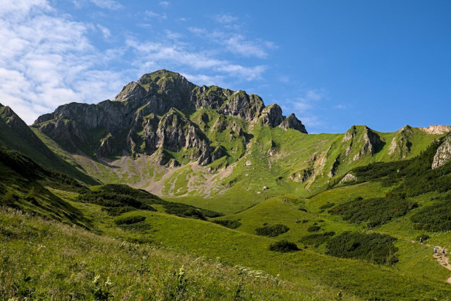 Grüne Almwiese im Vordergrund. Dahinter ragt ein Berg in die Höhe mit einem langgezogenen Kamm auf der rechten Seite. Es ist blauer Himmel mit etwas Wolken auf der linken oberen Seite.