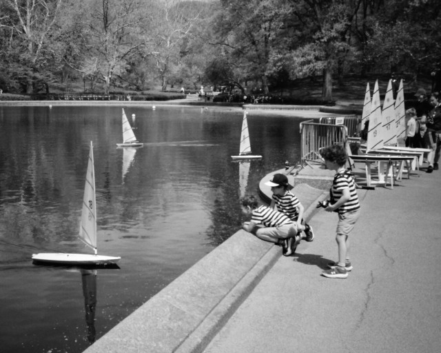 Three kids in matching striped shirts, one holding a remote control console, enjoy renting and racing one of Central Park's model Laser sailboats.