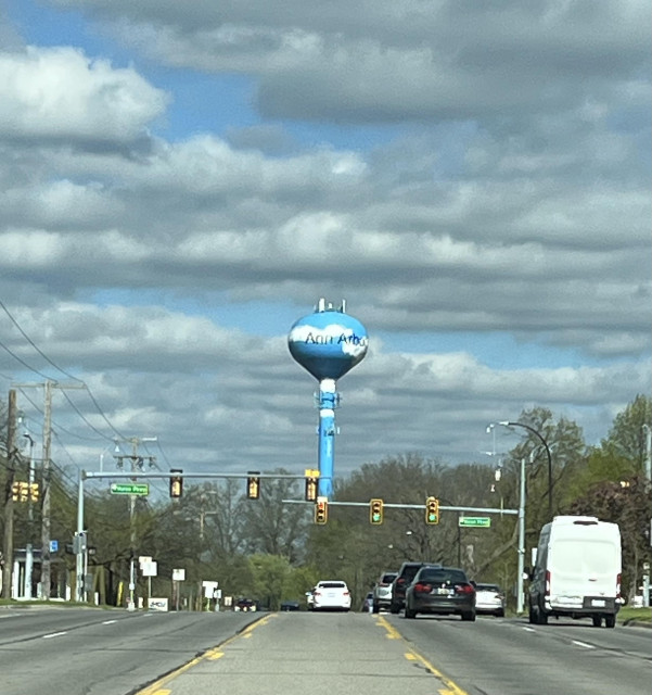 A late afternoon photo of a street scene. In the distance is a water tower painted sky blue with white fluffy clouds, and the name Ann Arbor painted in black lettering on its bulbous top. In the background is the sky that is blue with white/grey fluffy clouds. Photo taken on Plymouth Rd. off of Nixon Rd. in Ann Arbor, Michigan.