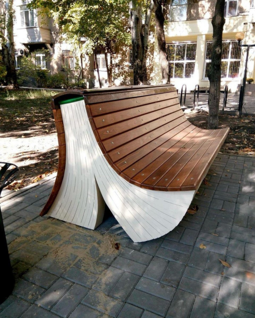 A photograph of a wooden park bench designed to look like an open, oversized book. The "pages" form the base and sides of the bench, while the wooden slats of the seat and back resemble the book’s cover. It is situated on a paved path in a park with trees and buildings in the background.