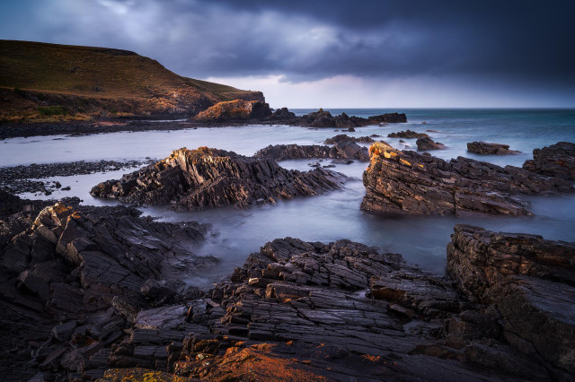 rugged coastal scene with jagged rock formations stretching diagonally through the frame, leading the eye toward the sea. The water has that smooth, misty look which contrasts nicely with the sharp textures of the rocks. In the background, there’s a sloping grassy cliff on the left, and the horizon sits under a heavy, moody sky with thick clouds rolling in.