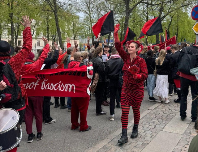 Ett demonstrationståg med rödsvarta fanor sett snett bakifrån. I vänstra hörnet skymtar en trumma. Delar av en röd sidobanderoll med vit text: "mot fascism" syns. En person klädd i rödsvart (zebra)randig klädsel går åt motsatt håll och signalerar till tåget med ena handen, samtidigt som den håller ett rödsvart häfte i den andra.