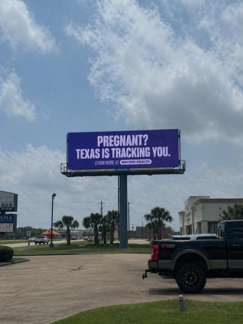 A large billboard displays the message "Pregnant? Texas is tracking you. Learn more at Mayday.Health" in front of a cloudy sky and near a parking lot.