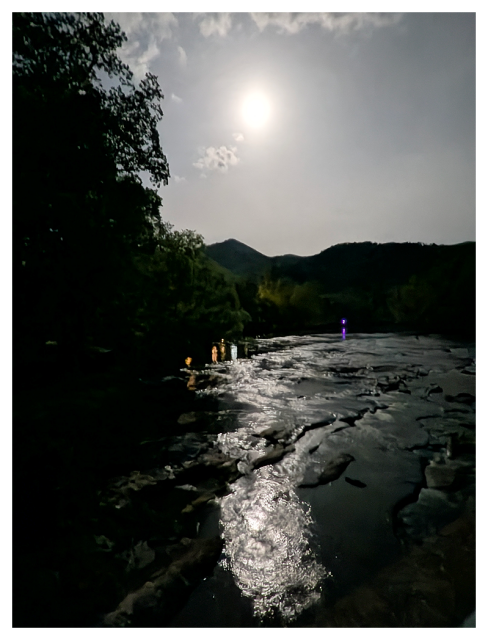 A bright full-appearing moon shines in a cloudy night sky over a rocky river, reflecting light on the water’s surface. Silhouetted trees and hills frame the scene. 