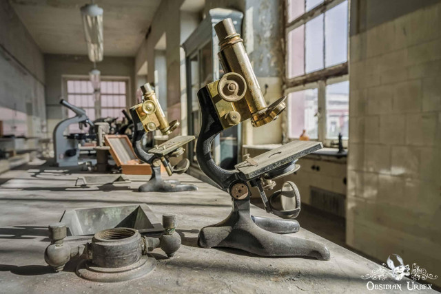 A vintage brass microscope sits on a dusty lab counter in an abandoned medical research institute, with other equipment