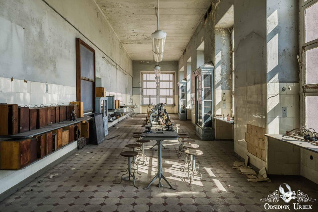 An abandoned medical teaching laboratory with peeling paint, long tables, stools, and vintage microscopes.