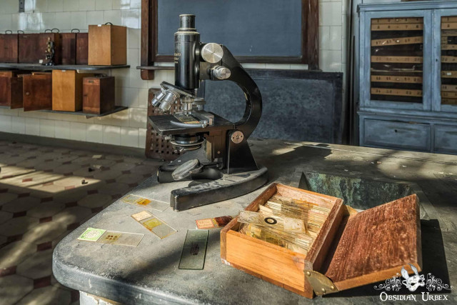 An antique microscope and open wooden box of slides sit on a dusty table in an abandoned lab.