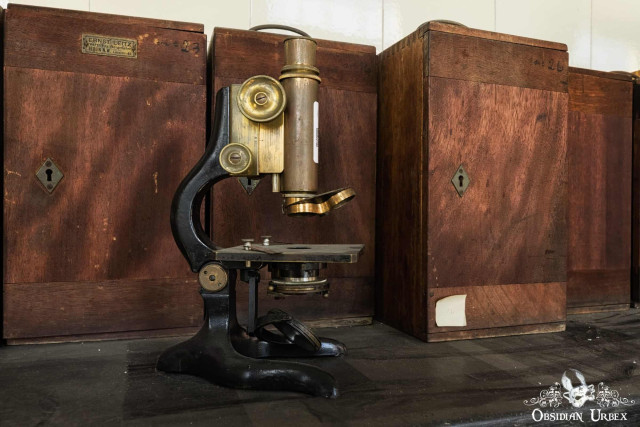 A vintage brass and black microscope stands on a dark surface before wooden storage boxes.