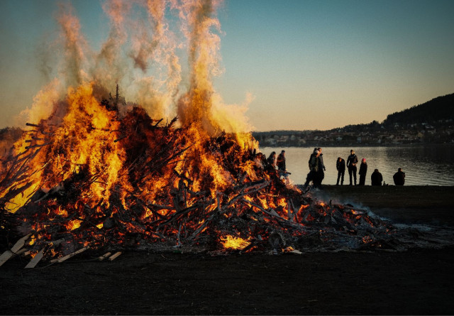 A massive bonfire blazes on the shoreline of Lake Storsjön at dusk, sending tall flames and smoke high into a pale blue sky. A group of people stand silhouetted against the lake to the right, watching the fire. Östersund’s cityscape and a forested hill are visible across the calm water in the background. The scene captures the traditional Swedish Walpurgis Night celebration in dramatic fashion