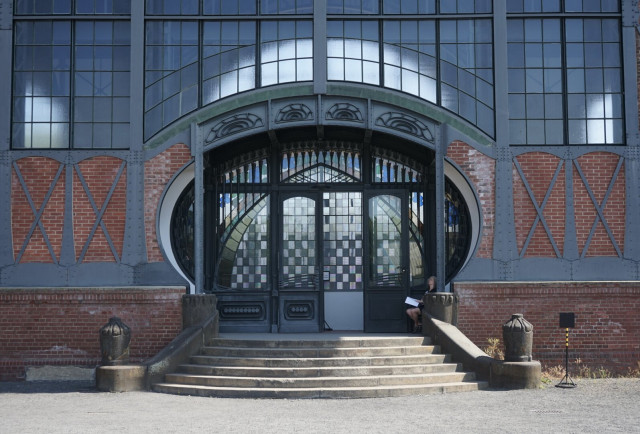 Dans une façade industrielle en brique, verre et métal, grande ouverture ovale en haut d’un escalier laissant voir des portes en vitraux décorées de damiers de couleurs.