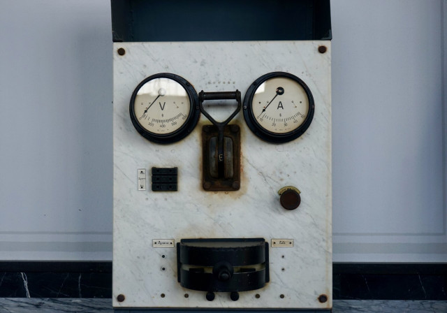 Petite machine blanche rectangulaire présentant un cadran à volts et un autre à ampères de part et d’autre d’un élément mal identifié, on a l’impression de voir deux yeux et un nez.