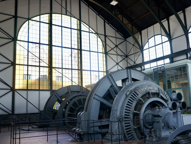 Machine d’extraction Siemens & Halske gris-bleu massive au pied d’une grande verrière bordée de vitraux jaunes qui laisse entrer une lumière généreuse.