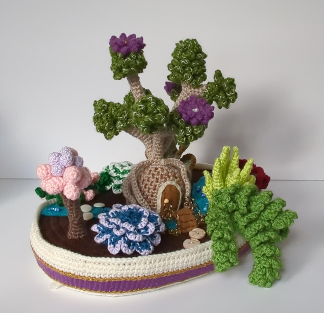 A crocheted planter containing crochet succulents. The planter is squat, wide and oval. A stream made of felt runs between the plants. The largest plant looks like a broccoli tree with purple flowers and a bulbous trunk. There's a rounded door on the front of the trunk lump and a small footbridge made of wire and felt just outside the door. The other plants are a variety of shapes, colors and heights: pastel pink and purple globes clustered on a brown stem; a dark aqua rosette with pale purple leaf edges; a medium green frilly trailing plant; and a yellow-green cluster of long pointy vertical leaves.