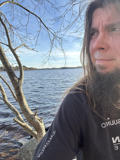 A man with long hair and a beard taking a selfie outdoors near a body of water, with a leafless tree and a clear sky in the background.