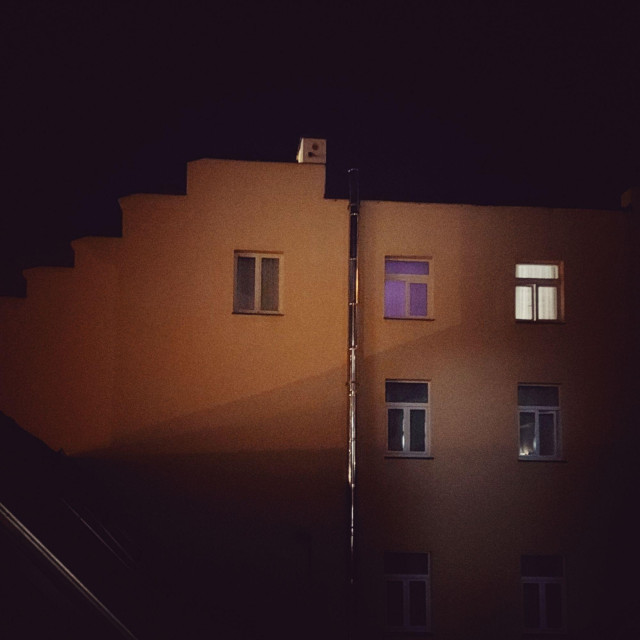 Wall of a house, dim against nightsky. Some windows, two of them lit.