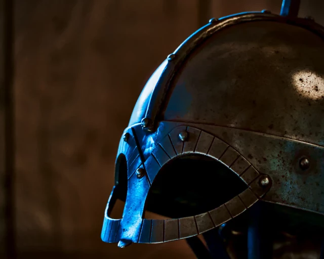 Der Helm eines Wikingers in einer Nahaufnahme. Nur der vordere Teil mit dem Augenschutz ist zu sehen. Der Helm steht auf einem Gerüst vor einer dunklen Lehmwand. Von links kommt irgendwoher Licht in den normalerweise dunklen Raum und lässt das Metall des Helmes blau aufleuchten. 