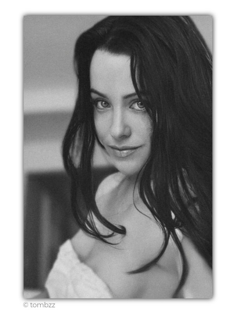 A black and white, close-up portrait of my wife, Elisa, captured through film photography. Elisa has long, dark, slightly wavy hair that falls softly over her bare shoulders. She is looking directly into the lens, her gaze incredibly deep, calm, and magnetic. A barely perceptible, tender smile plays on her lips. The image has a grainy texture and soft contrast, creating an intimate, timeless atmosphere. The background is softly blurred and non-distracting, keeping the focus entirely on Elisa's face and emotions.