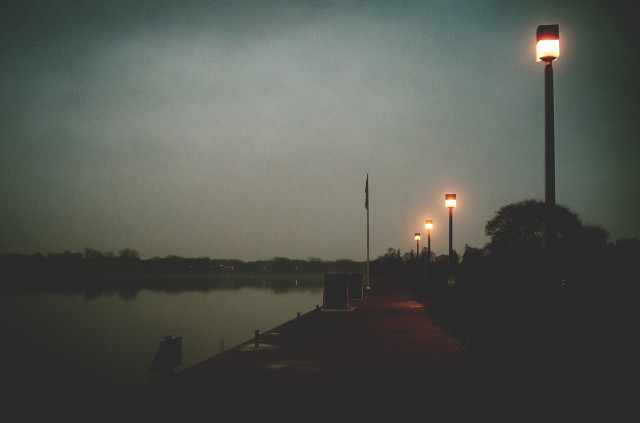 Early morning beside the calm waters of a lake. The photographer is standing on a cobblestone walkway beside the water, the glow of lights on lampposts are seen. The overall feel of the image is muted and dream like. 
