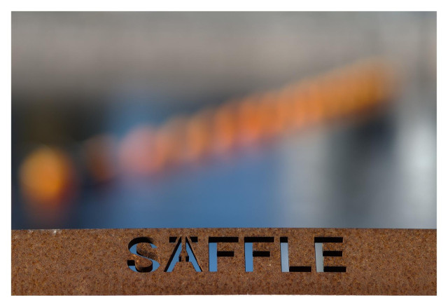 En liggande bild på ordet "SÄFFLE" urfräst ur en rostig metalskylt. Ovanför syns en rad av orangea bojar i blått vatten i kraftig oskärpa.