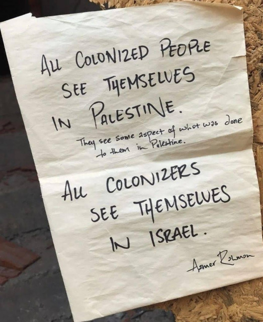 A photograph of a piece of white paper with black handwritten text, attached to a textured, tan-colored surface. The top of the paper reads in large, capitalized letters, "ALL COLONIZED PEOPLE SEE THEMSELVES IN PALESTINE." Below this, smaller text reads, "They see some aspect of what was done to them in Palestine." The lower section of the paper states in large, capitalized letters, "ALL COLONIZERS SEE THEMSELVES IN ISRAEL." At the bottom right, there is a signature that reads, "Amer Rahman."