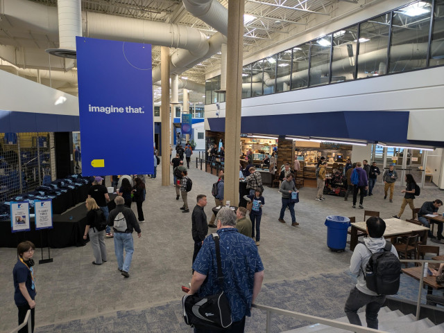 A picture atop a flight of stairs extending down a lengthy, carpeted corporate campus corridor. People mill about. All the accents are bright blue.