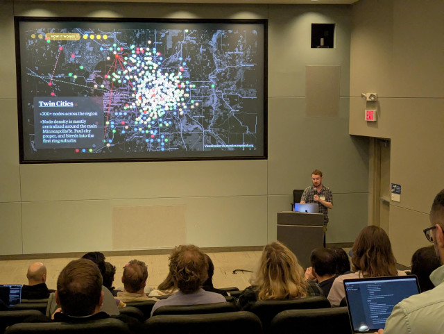 A speaker stands in front of a theater of people. On the screen is a dark mode map of Minneapolis/St Paul. It is covered with dots representing mesh nodes.