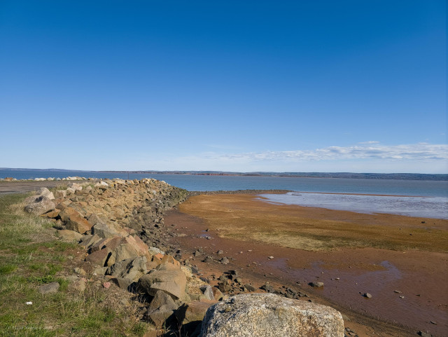 A scene looking over the bay with a rocky shoreline curving around a wet beach. The sky is blue with hardly any clouds.