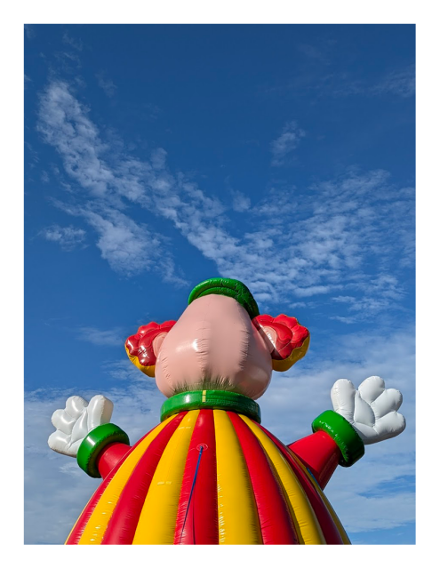 an inflatable clown, seen from behind, wears a red and yellow striped outfit with green trim and white gloves. Its head is large with red ears or hair. the clown stands against a bright blue sky with wispy clouds.
