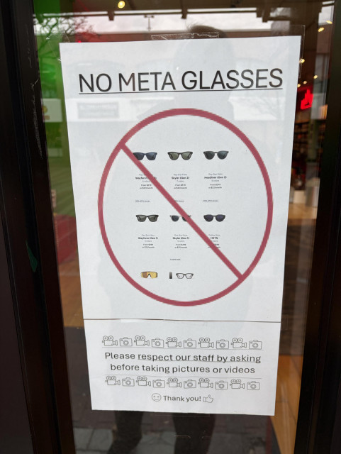 A sign on a store window indicates "NO META GLASSES," featuring images of different sunglasses models. It also includes instructions to respect staff by asking permission before taking pictures or videos, with a friendly "Thank you!" at the bottom.