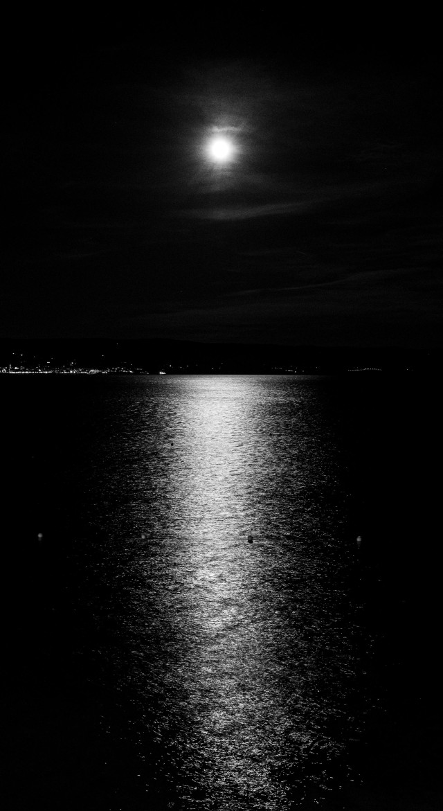 Black and white photo of the full moon with a few light clouds reflecting on the sea. High contrast so all you can see is the moon, reflections, lights of a distant town, and traces of some clouds.